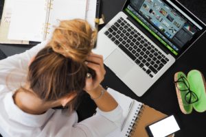 woman with bun, hand on head, laptop, phone and notebooks