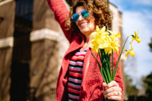 woman in sunglasses, red leather jacket, and striped dress holding yellow flowers