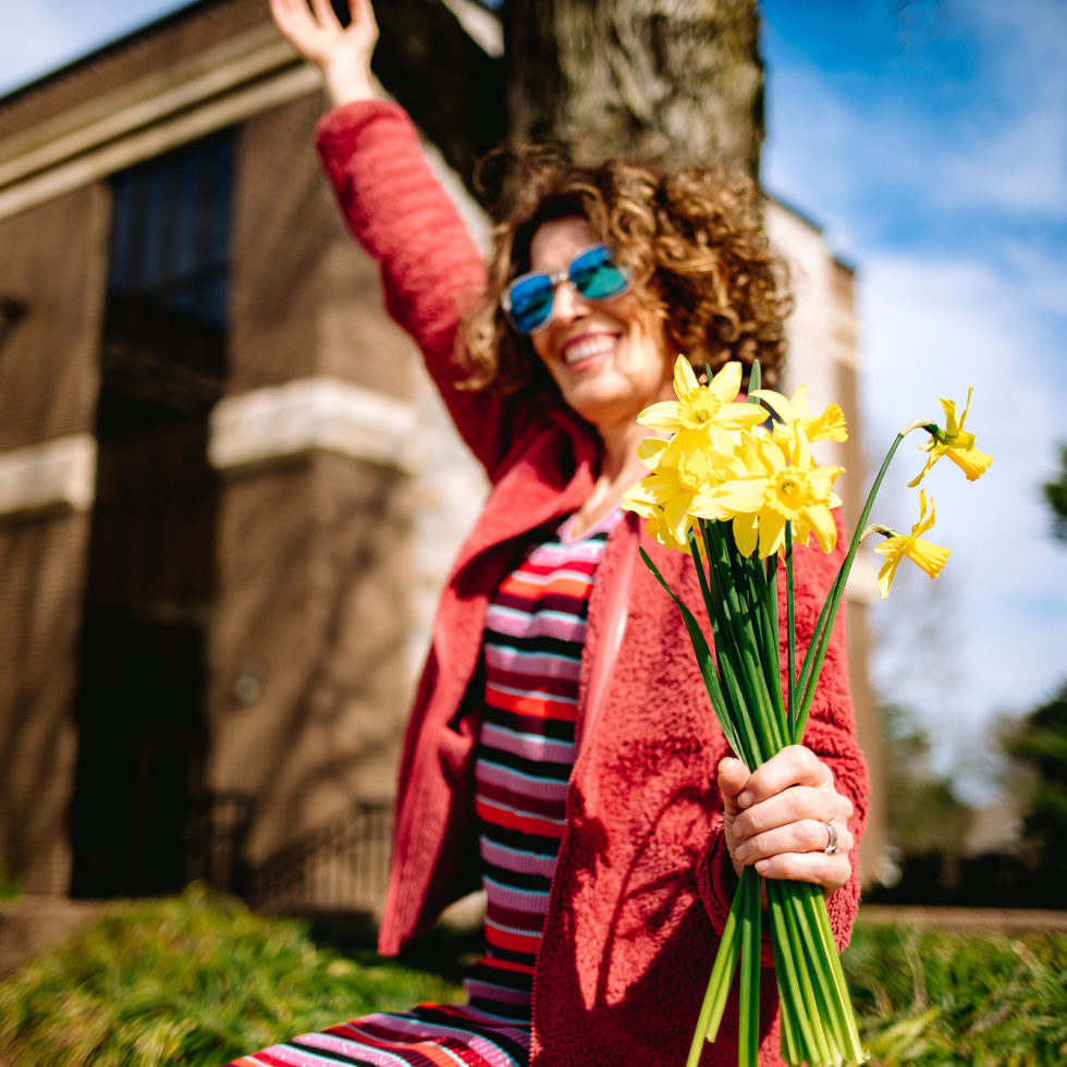 woman in sunglasses, red leather jacket, and striped dress holding yellow flowers